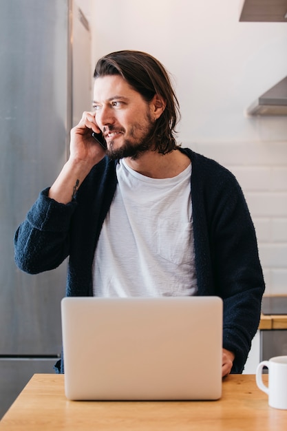 Gratis foto portret van een knappe jonge man praten op mobiele telefoon met laptop op tafel