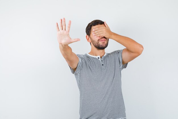 Portret van een knappe man die zijn ogen bedekt met de hand, een stopgebaar in een t-shirt laat zien en een beschaamd vooraanzicht toont