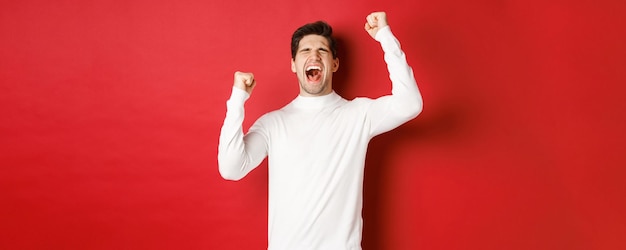 Portret van een knappe man in een witte trui, die zich vrolijk voelt, de overwinning viert, schreeuwt van vreugde en de handen opsteekt in de overwinning, staande op een rode achtergrond.