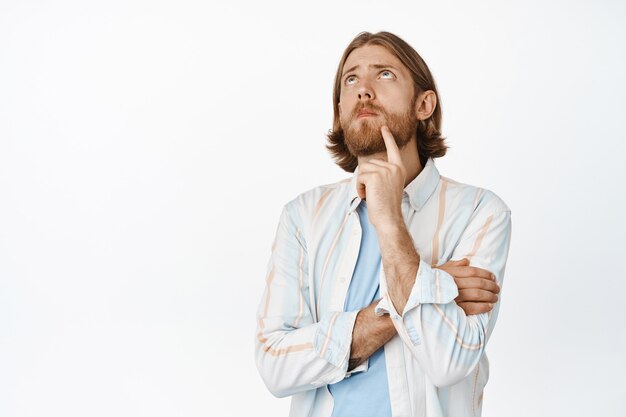 Portret van een knappe nadenkende blonde man, die zijn baard aanraakt, fronst en opkijkt, aan iets interessants denkt, peinzend, staande op wit.