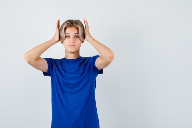 Portret van een knappe tienerjongen die de handen op het hoofd houdt in een blauw t-shirt en er opgewonden uitziet