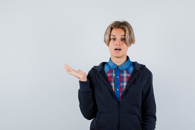 Portret van een knappe tienerjongen die de palm opzij spreidt in hemd, hoodie en een verbaasd vooraanzicht kijkt