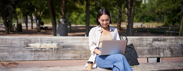 Gratis foto portret van een koreaanse vrouw die met een laptop in het park op een bank zit en buitenshuis werkt