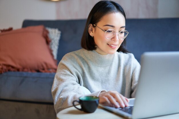 Portret van een lachend Aziatisch meisje dat vanuit huis werkt en op afstand blijft met behulp van een laptop die op haar computer studeert