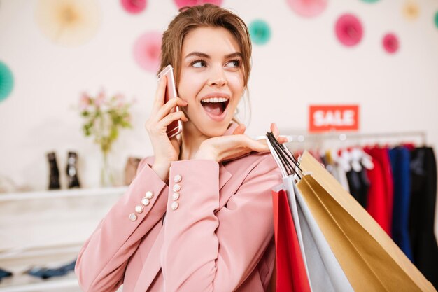 Portret van een lachend meisje in een roze jas die gelukkig opzij kijkt met kleurrijke boodschappentassen op haar schouder en mobiele telefoon in de hand in de kledingwinkel. Jonge dame praat op haar mobiel in boetiek