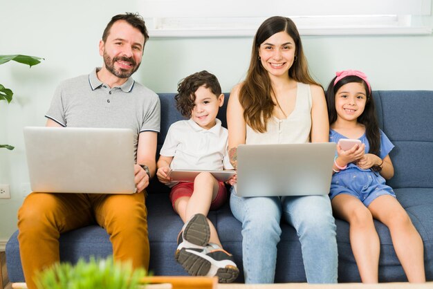 Portret van een lachende jonge moeder en Latijnse vader die laptops vasthoudt met hun kleine kinderen, zit met een tablet en een smartphone in de woonkamer