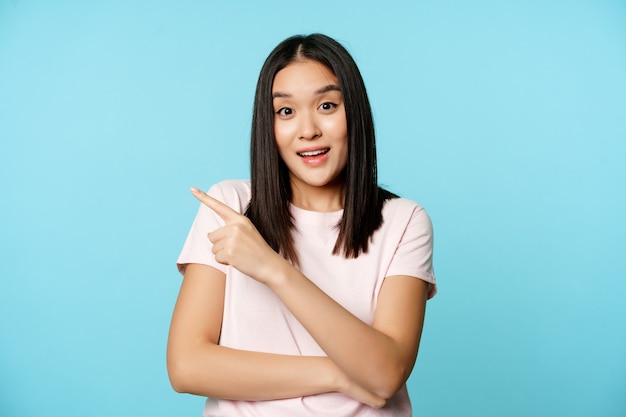 Portret van een lachende Koreaanse vrouw die naar de linkerbovenhoek wijst, met korting, verkoopbanner, in een t-shirt op een blauwe achtergrond