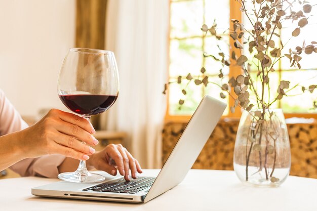 Portret van een lachende volwassen senior vrouw die een glas wijn vasthoudt terwijl ze een laptop op de keukentafel gebruikt. freelance thuiswerken concept.