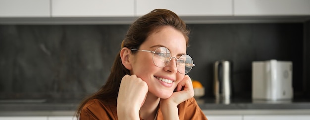 Gratis foto portret van een lieve gelukkige jonge vrouw met een bril die in de keuken zit met een dromerig bedachtzaam gezicht