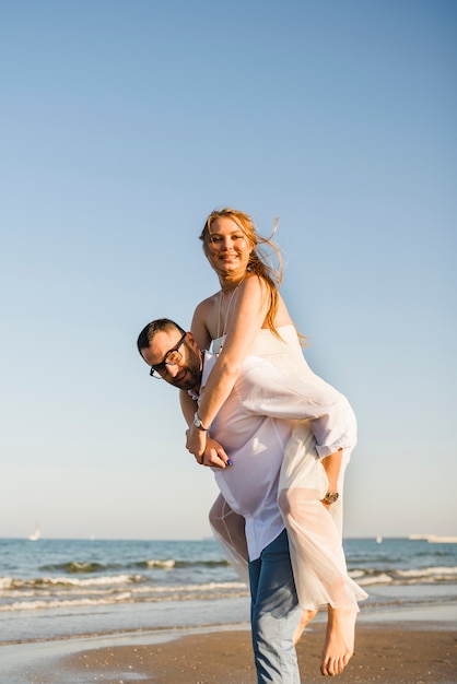 Portret van een man die op de rug rit geeft aan haar vrolijke vrouw op strand tegen blauwe heldere hemel