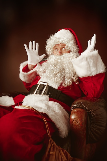 Portret van een man in kerstman-kostuum - met een luxe witte baard, kerstmuts en een rood kostuum op een rode studioachtergrond