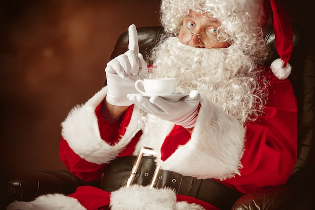 Portret van een man in kerstman kostuum met een luxe witte baard, kerstmuts en een rood kostuum op rood zittend in een stoel met een kopje koffie