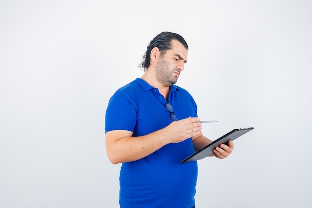 Portret van een man van middelbare leeftijd die door klembord kijkt terwijl hij potlood in polot-shirt vasthoudt en nadenkend vooraanzicht kijkt