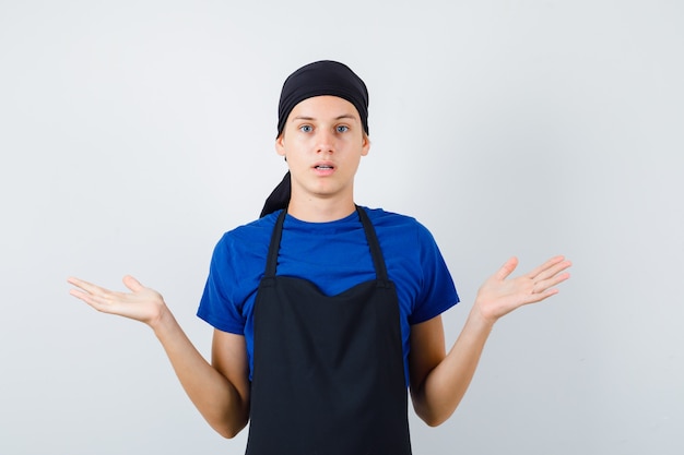 Gratis foto portret van een mannelijke tienerkok die een hulpeloos gebaar toont in een t-shirt, een schort en een aarzelend vooraanzicht