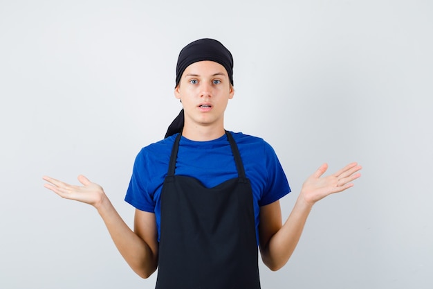 Portret van een mannelijke tienerkok die een hulpeloos gebaar toont in een t-shirt, een schort en een aarzelend vooraanzicht