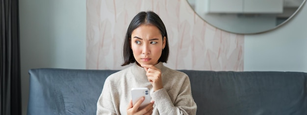 Gratis foto portret van een meisje dat op de bank zit en met een smartphone nadenkend en aarzelend naar de mobiele telefoon kijkt