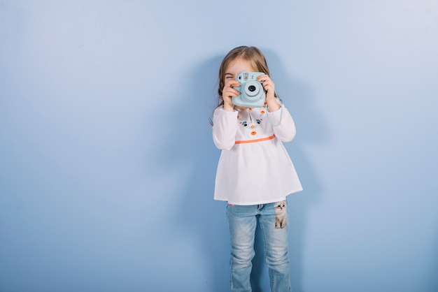 Portret van een meisje die foto met uitstekende onmiddellijke camera nemen die zich tegen blauwe achtergrond bevinden