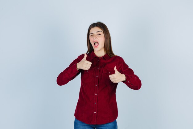 Portret van een meisje met dubbele duimen omhoog in bordeauxrood shirt en er geamuseerd uitziet
