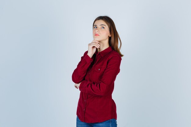 Portret van een meisje met haar kin in haar hand, omhoog kijkend in bordeauxrood shirt en bedachtzaam kijkend