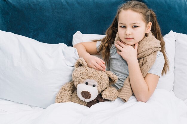 Portret van een meisje wat betreft haar hals met hand die met teddybeer op bed situeert