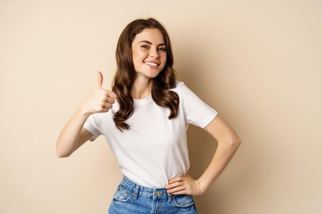Portret van een moderne jonge vrouw die duimen laat zien, leuk vindt en goedkeurt, tevreden glimlacht, bedrijf of website aanbeveelt, staande over een beige achtergrond.