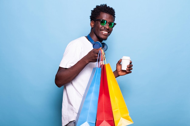 Portret van een moderne man met boodschappentassen en koffiedrank in de hand. zorgeloze persoon die een zonnebril draagt en glimlacht terwijl hij handtassen draagt na het kopen van kleding in de uitverkoop. winkelplezier