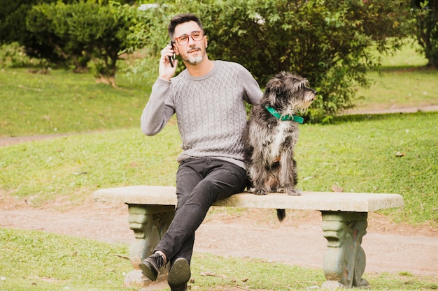 Gratis foto portret van een moderne man zit in park met zijn hond praten op mobiele telefoon