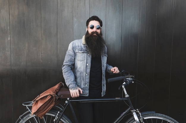 Portret van een modieuze jonge mens die zonnebril draagt die zich voor zwarte houten muur met de fiets bevindt