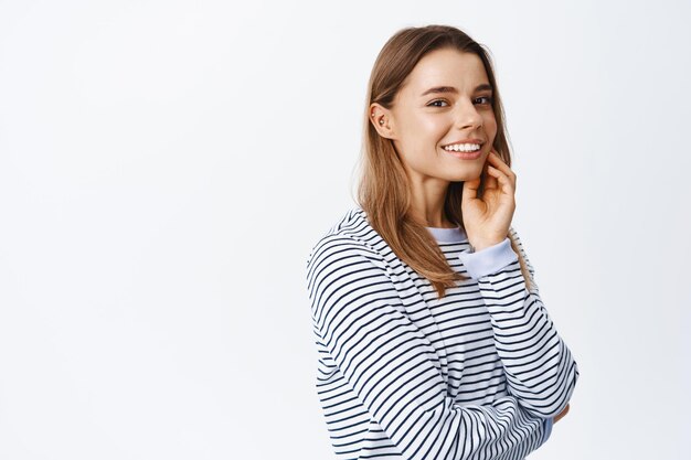 Portret van een mooi blond meisje dat haar gezicht aanraakt en oprecht glimlacht, een zuivere, stralende huid laat zien zonder make-up, over wit staat