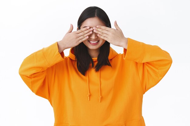 Portret van een mooi jong Aziatisch meisje in een oranje hoodie, sluit de ogen met handpalmen en lacht geamuseerd, viert b-day in afwachting van een verrassingscadeau, speelt verstoppertje, staande witte muur