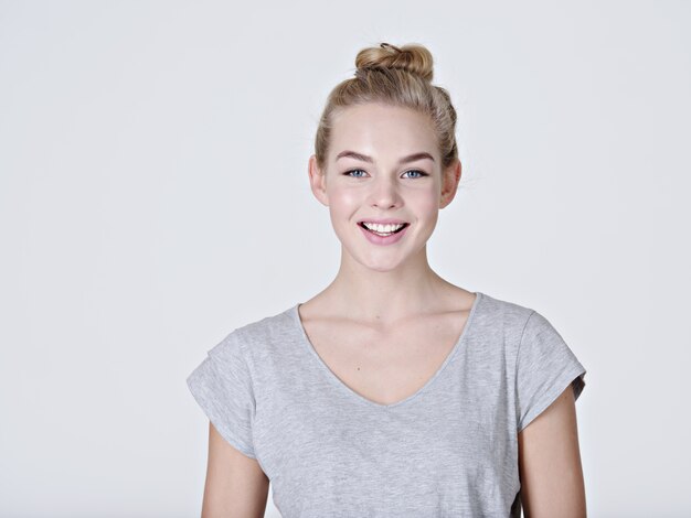 Portret van een mooi jong lachend meisje. Vrouwelijk gezicht met brede glimlach. Aantrekkelijk blond meisje met zich meebrengt in de studio in een casual grijs t-shirt