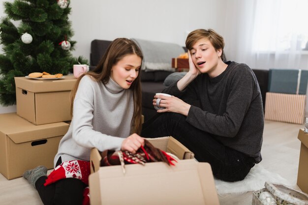 Portret van een mooi jong stel dat thuis op de vloer zit en verbaasd in een doos kijkt met een kerstboom op de achtergrond