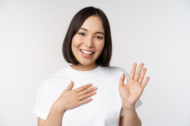 Portret van een mooi Koreaans meisje dat de hand opsteekt, stelt zichzelf voor en legt de hand op een hartgroet die op een witte achtergrond staat