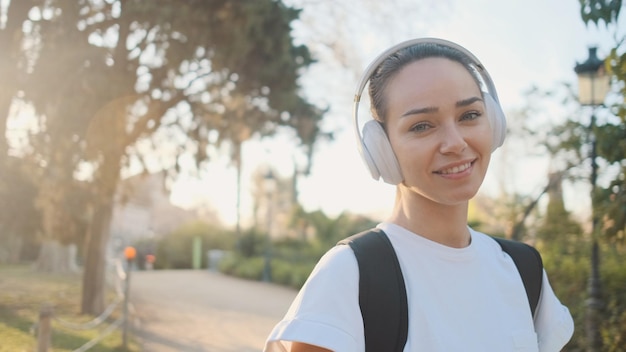 Portret van een mooi meisje in een koptelefoon met een rugzak die lacht op de camera op weg naar de ochtendtraining in het stadspark