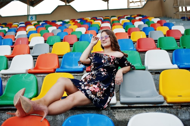 Gratis foto portret van een mooi meisje in jurk en zonnebril zittend op de tribunes in het stadion