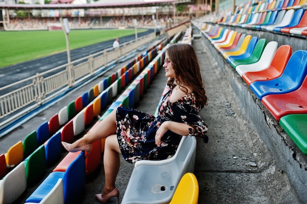 Portret van een mooi meisje in jurk en zonnebril zittend op de tribunes in het stadion