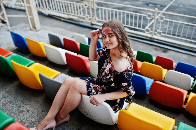 Portret van een mooi meisje in jurk en zonnebril zittend op de tribunes in het stadion