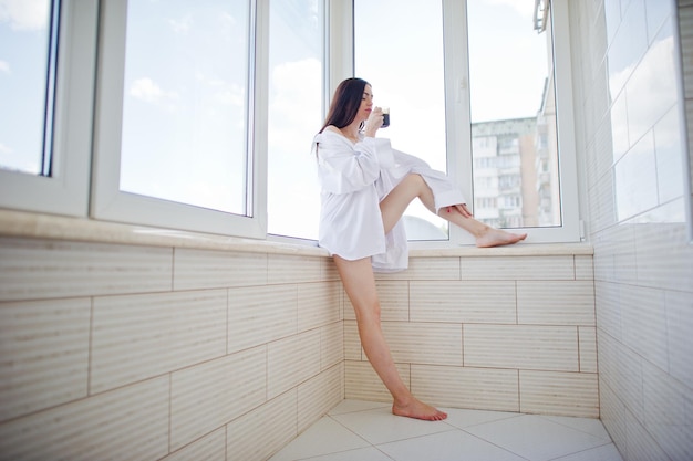 Portret van een mooi meisje in ondergoed en mannelijk overhemd dat met een glas water in haar handen op het balkon staat