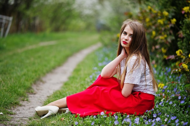 Portret van een mooi meisje met rode lippen in de lentebloesemtuin op gras met bloemen op een rode jurk en een witte blouse