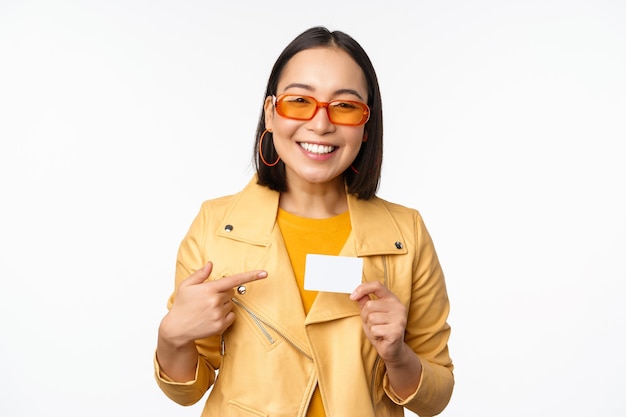 Portret van een mooi modern Aziatisch meisje in een zonnebril die lacht en blij is met een creditcard die over een witte achtergrondkopieerruimte staat