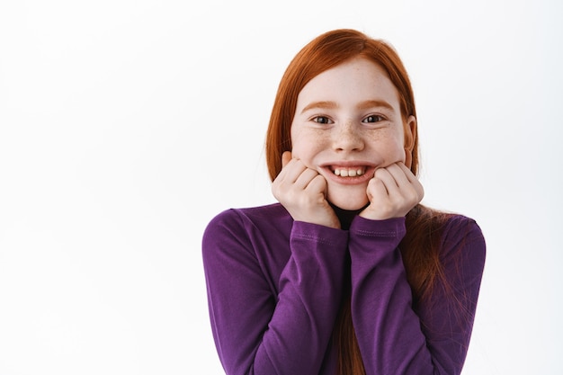 Portret van een mooi roodharig kind, een klein gembermeisje met sproeten raakt de wangen aan en glimlacht gelukkig aan de voorkant, ziet er schattig en schattig uit, witte muur