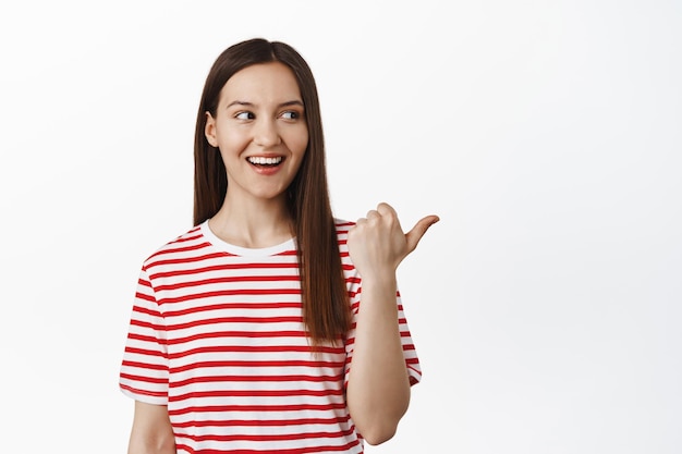 Portret van een mooie 20s-vrouw die met de vinger naar rechts wijst, glimlacht en naar het logo kijkt, vertelt over een groot verkoopevenement, kortingen in de winkel, staande tegen een witte achtergrond