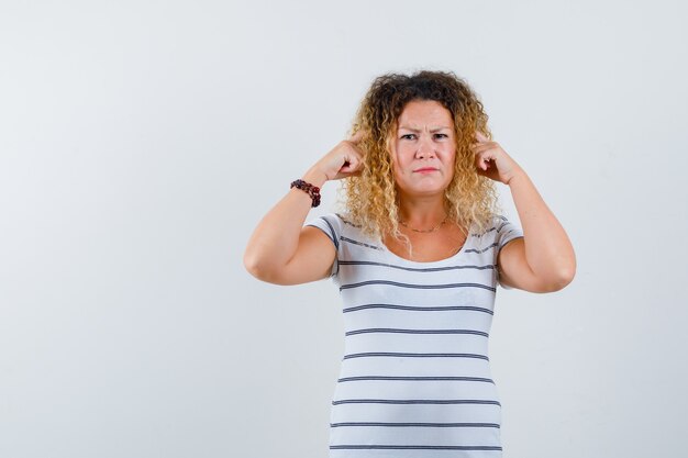 Portret van een mooie blonde vrouw die vingers op de slapen houdt in een gestreept t-shirt en er ontevreden vooraanzicht uitziet