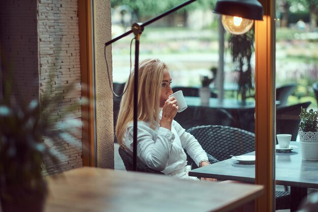 Portret van een mooie blonde vrouw gekleed in een witte blouse zittend aan een tafel drinkt koffie op terras café.