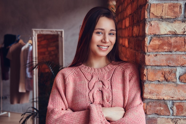 Portret van een mooie charmante brunette gekleed in stijlvolle kleding glimlachend terwijl ze tegen een bakstenen muur leunt in een kamer met loft-interieur.