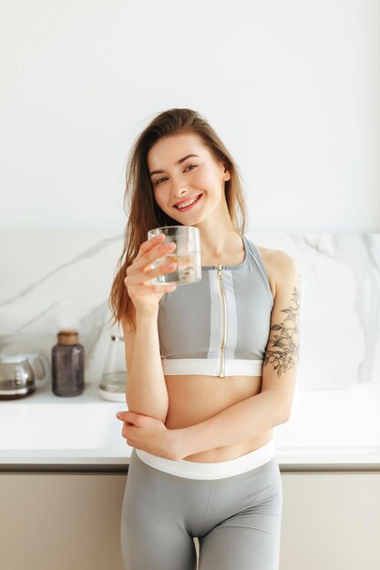 Portret van een mooie dame in een sportieve top die op de keuken staat en vrolijk in de camera kijkt met een glas water