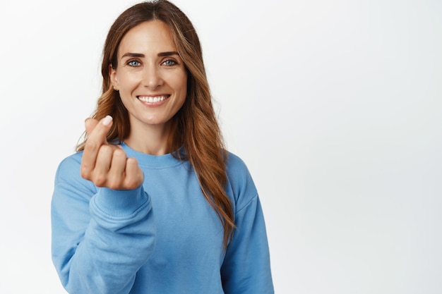 Portret van een mooie en gelukkige vrouw die lacht, met een hartgebaar van de vinger, staande in vrijetijdskleding tegen een witte achtergrond