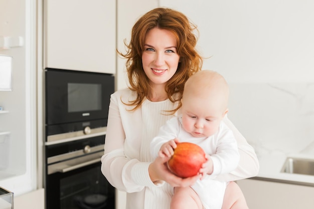 Portret van een mooie glimlachende moeder die in de keuken staat en gelukkig in de camera kijkt terwijl ze haar schattige kleine baby en grote rode appel in handen houdt