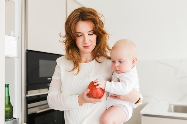 Portret van een mooie glimlachende moeder die in de keuken staat en haar schattige kleine baby en grote rode appel in handen houdt