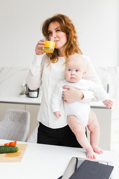 Portret van een mooie glimlachende vrouw die haar schattige kleine baby vasthoudt terwijl ze sap drinkt en kookt in de keuken